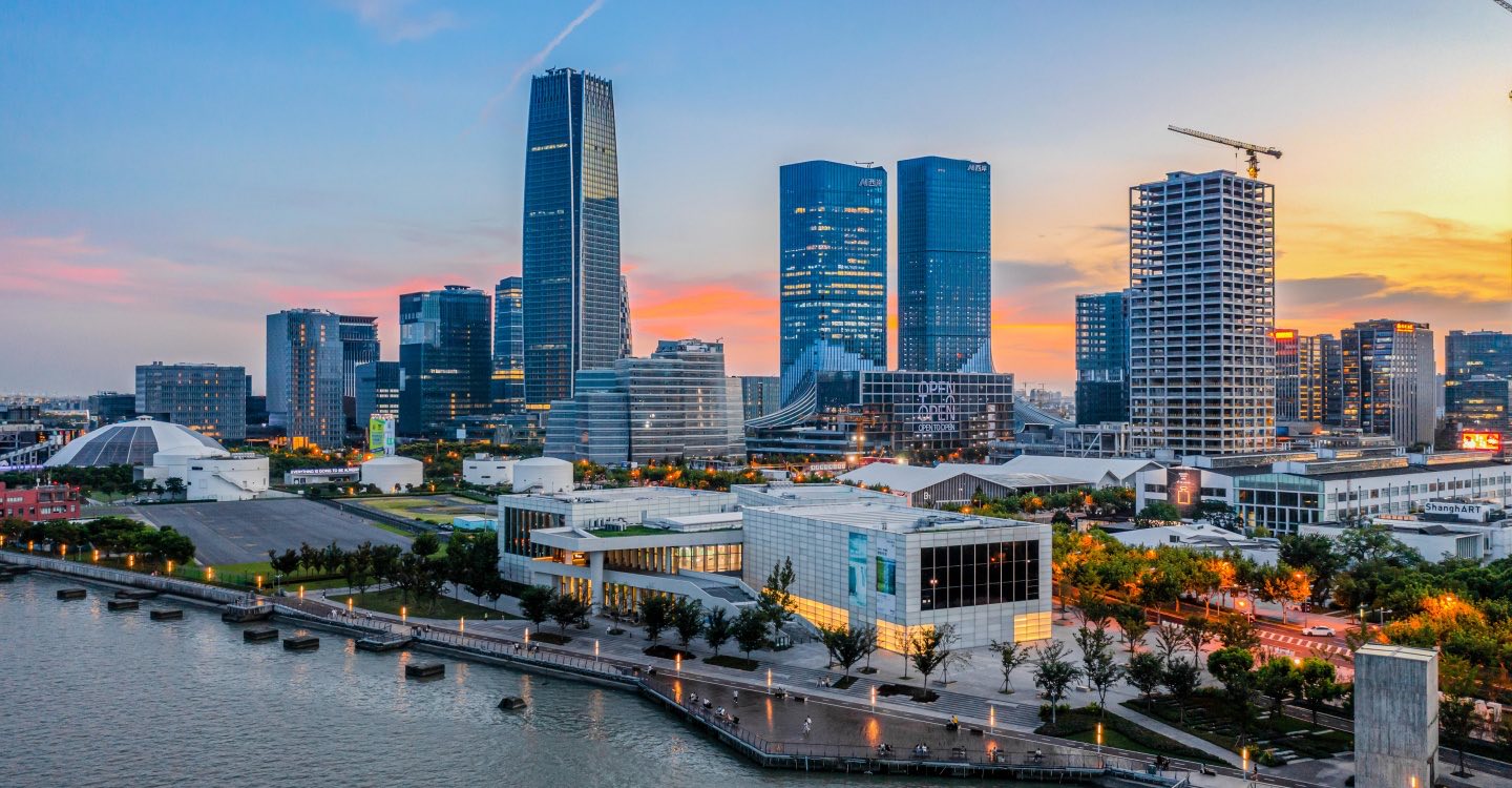 The West Bund riverfront in Xuhui, Shanghai, at dusk: towers along the Huangpu behind a low glass museum and a planted promenade.